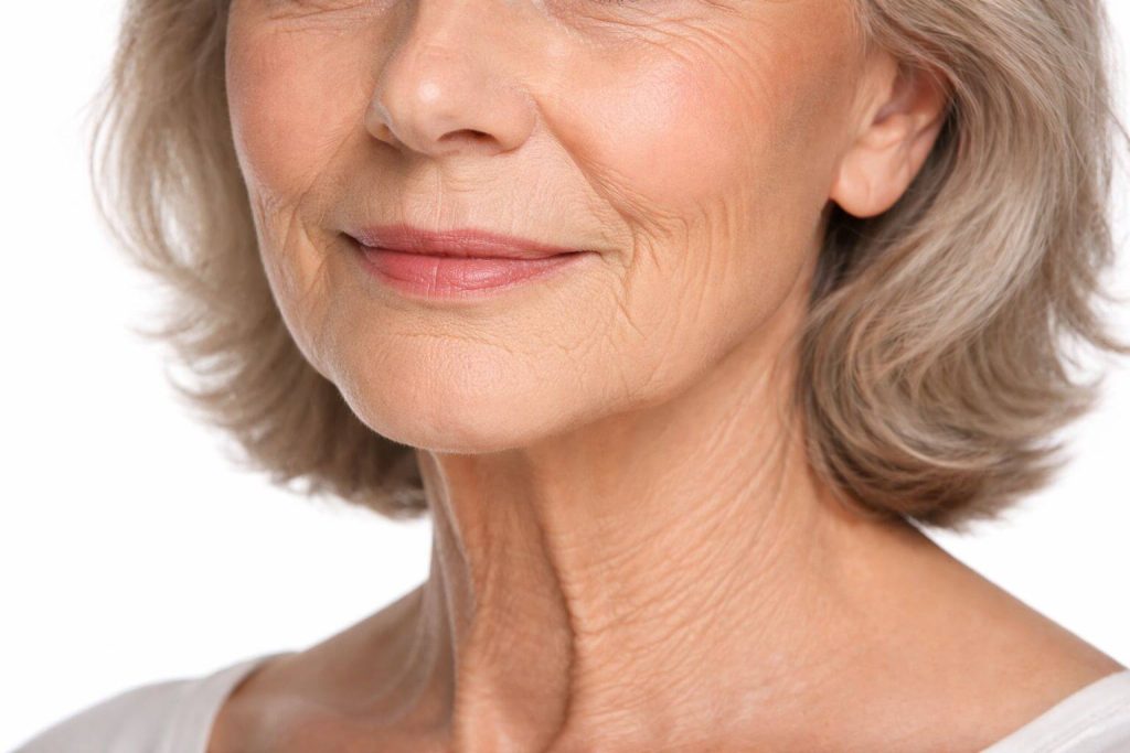 Uśmiechnięta starsza kobieta z siwymi włosami, ukazująca zmarszczki na twarzy i szyi, na jasnym tle.